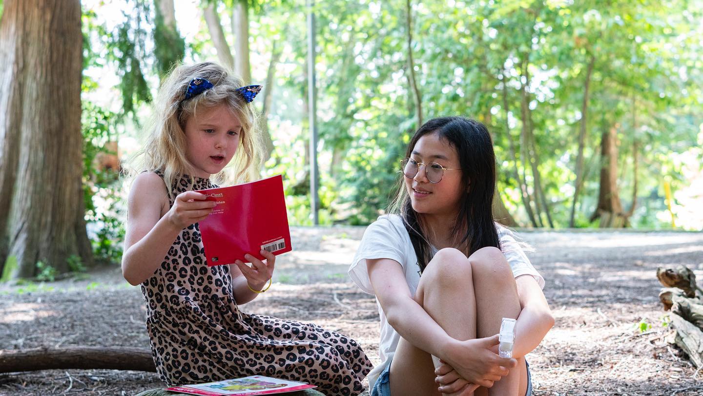 Young student reading a book to another student while sitting on a forest path